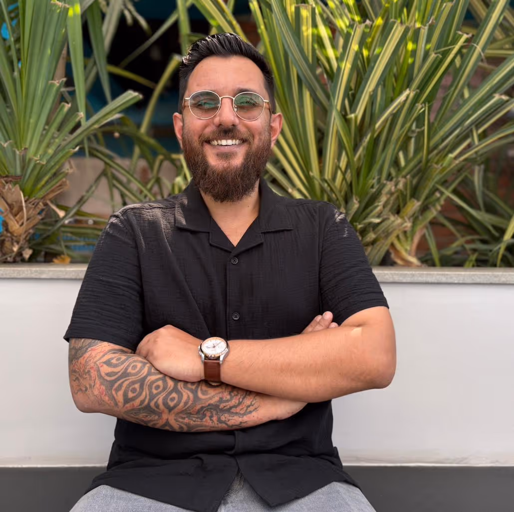 Smiling man with glasses, beard, and tattooed arm crossing his arms, sitting in front of green plants.