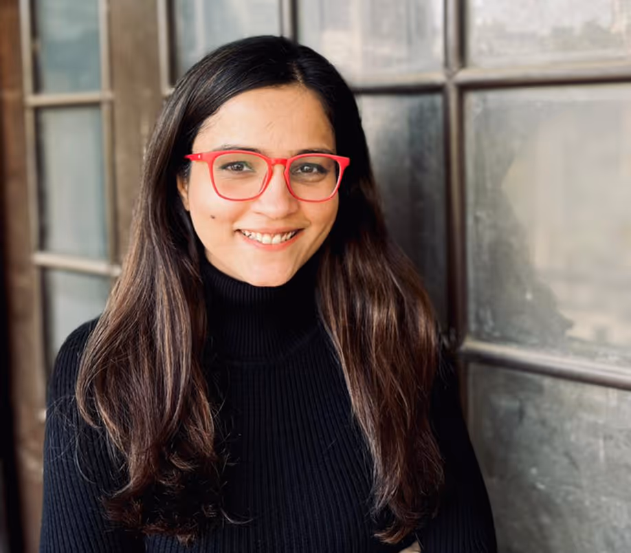 Woman with long dark hair wearing red glasses and a black turtleneck sweater smiling in front of a windowed wall.